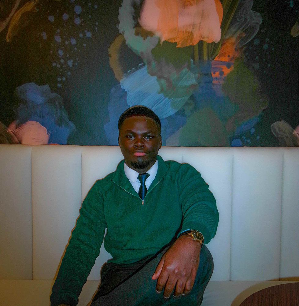 Isaac Adjei in a green sweater and tie, seated on a white bench in front of a colorful abstract mural